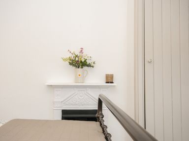 A beige bedspread on a metal bed frame next to a white mantel with a vase of flowers and a candle on it at Skylarks in No Mans Land near Looe