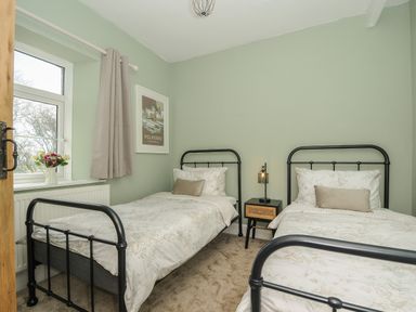 A bedroom with two single beds a nightstand with a lamp a window with curtains and a framed picture on the wall at Skylarks in No Man's Land near Looe