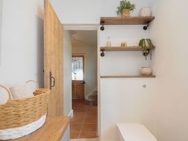 A bathroom with wooden shelves holding plants and candles and a wooden door leading to a hallway with tiled floor at Skylarks in No Mans Land near Looe