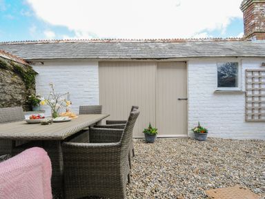 An outdoor patio with a wicker dining table and chairs next to a white brick wall with a wooden door and window at Skylarks in No Man's Land near Looe