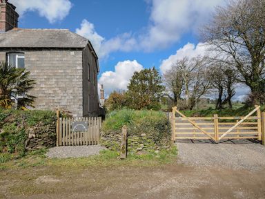 A stone house with a wooden fence and gate next to a gravel driveway and trees at Skylarks in No Mans Land near Looe