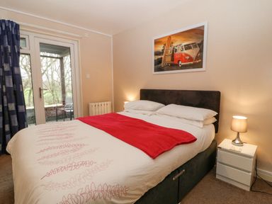 A bedroom with a bed covered with white and red bedding a bedside table with a lamp and glass doors leading outside at No 51 Valley Lodges in Honicombe Manor near Gunnislake