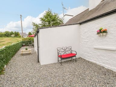A pebble patio with a metal bench with a red cushion a wooden picnic table and a flower pot on a white cottage exterior at Cartron Cottage in Ballintubber County Mayo