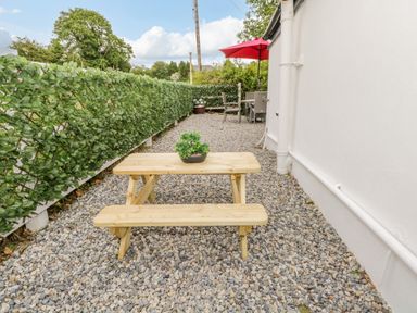 An outdoor area with a wooden picnic table and benches on gravel next to a white wall and green hedge at Cartron Cottage in Ballintubber County Mayo