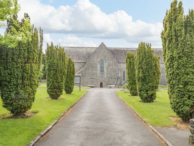 A paved path lined with tall trees leading to a stone building with arched windows at Cartron Cottage in Ballintubber County Mayo