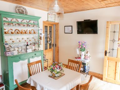 A dining room with a table and chairs a green cabinet with dishes a wall mounted TV and flowers on a side table at Cartron Cottage in Ballintubber County Mayo