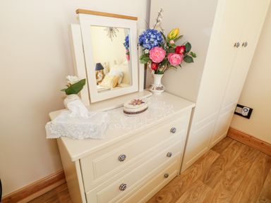 A white dresser with a mirror flowers and a lace tissue box on top in a room with wooden flooring at Cartron Cottage in Ballintubber County Mayo