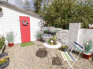 An outdoor seating area with a small table, two chairs, flower pots, and a red door on a white shed at Cartron Cottage in Ballintubber County Mayo