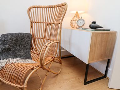 A rattan chair with a dark patterned blanket next to a wooden sideboard with a lamp clock and bird figurine at Stoker's Rest in Whitby