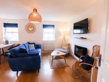 A living room with a blue sofa a patterned chair a rattan chair a coffee table a wall-mounted tv and two windows at Stoker's Rest in Whitby