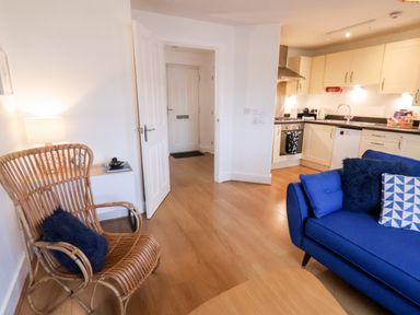 A living room with a blue sofa and a wicker chair next to an open door leading to a kitchen at Stoker's Rest in Whitby