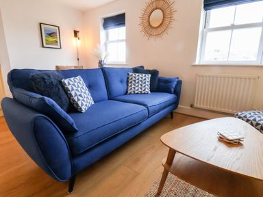 A living room with a blue sofa including patterned cushions a wooden coffee table and windows at Stoker's Rest in Whitby