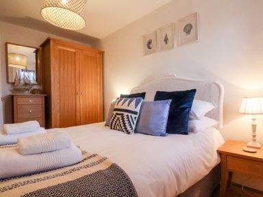 A bedroom with a bed, multiple pillows, framed shell pictures on the wall, a wooden wardrobe, chest of drawers, and a bedside table with a lamp at Stoker's Rest in Whitby