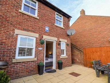 An exterior view of a brick house with white framed windows an open door leading inside two black planters with green plants a satellite dish a wooden fence and a patio area with outdoor seating at Stoker's Rest in Whitby