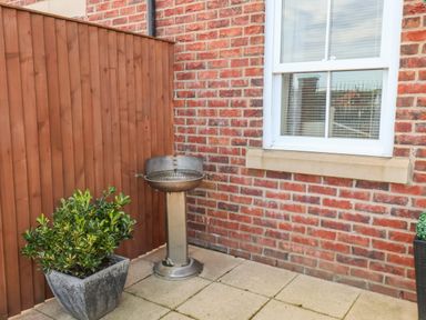 A patio area with a plant in a square pot a metal barbecue grill and a window on a brick wall at Stoker's Rest in Whitby