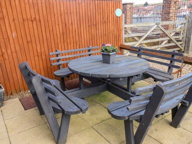 An outdoor seating area with a round wooden table and attached benches with a flower pot on top at Stoker's Rest in Whitby