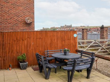 An outdoor patio with a round wooden table and attached benches a potted plant on the table a wooden fence and brick wall in the background at Stoker's Rest in Whitby