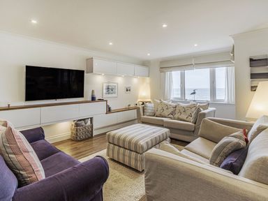 A living room with sofas a striped ottoman a wall-mounted television and a window overlooking the sea at Apartment 18 Burgh Island Causeway in Bigbury-On-Sea