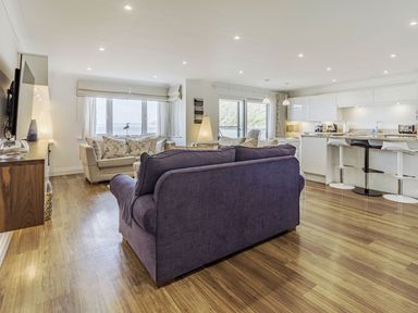 An open living room with a purple sofa and beige chairs facing a wall-mounted TV with a floating media console and a kitchen with white cabinets and bar stools at Apartment 18 Burgh Island Causeway in Bigbury-On-Sea