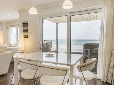 A dining area with white chairs and a table by glass doors overlooking a balcony with wicker chairs and the sea beyond at Apartment 18 Burgh Island Causeway in Bigbury-On-Sea