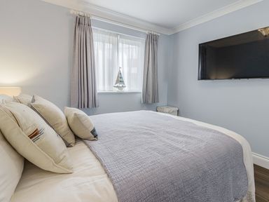 A bedroom with a bed pillows curtains a small sailboat decoration on the windowsill a wall mounted television and a small cabinet at Apartment 18 Burgh Island Causeway Bigbury-On-Sea