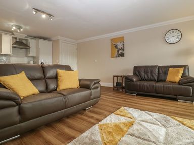 A living room with brown leather sofas and yellow cushions a wooden floor a clock on the wall and a kitchen area at Townbridge Apartment in Weymouth
