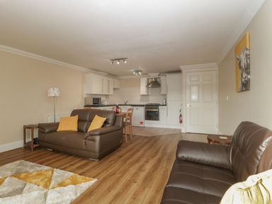 An open living room and kitchen area with brown sofas yellow cushions a wooden dining table and white kitchen cabinets at Townbridge Apartment in Weymouth