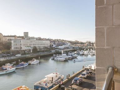 A marina with multiple boats docked next to a waterfront road with parked cars and buildings in the background at Townbridge Apartment in Weymouth