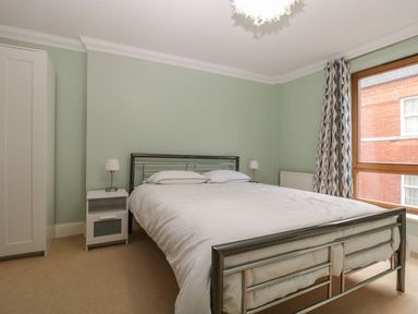 A bedroom with a metal bed frame white bedding two bedside tables with lamps a white wardrobe and a window with patterned curtains at Townbridge Apartment in Weymouth