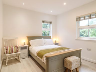 A bedroom with a bed a rocking chair a nightstand with a lamp and two windows at Dreamwood Lodge in Keston near Leaves Green