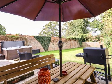 A wooden picnic table with a large umbrella and a barbecue grill in a garden with a hot tub and chairs at Dreamwood Lodge in Keston near Leaves Green