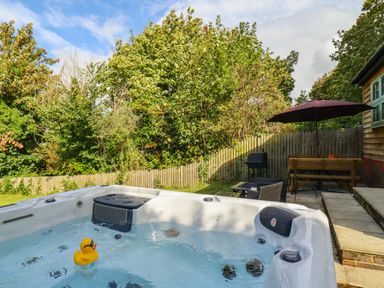 An outdoor hot tub with a yellow rubber duck in a garden next to a patio with table chairs umbrella and barbecue at Dreamwood Lodge in Keston near Leaves Green