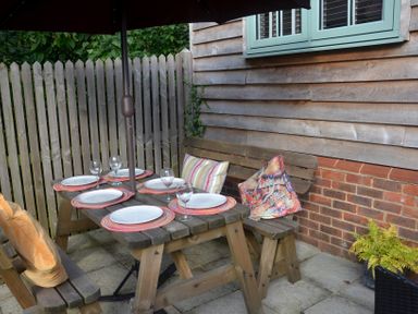 An outdoor wooden picnic table set with plates and wine glasses next to a wooden fence and house wall at Dreamwood Lodge in Keston near Leaves Green