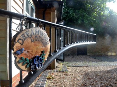 A wooden sign reading Dreamwood Lodge hanging on a black metal railing outside a building with gravel and brick pavement at Dreamwood Lodge Keston near Leaves Green