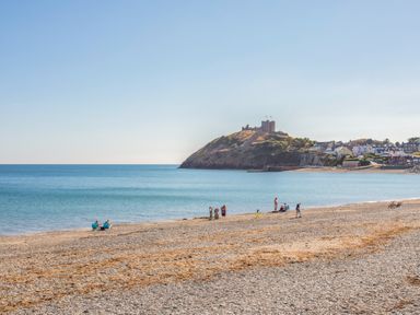 A pebble beach with people near the water and a hill with a castle and houses in the background at The Hideaway Pod in Rhoslan near Criccieth