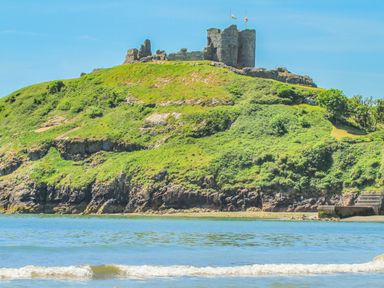 A hill with ruins of a stone castle and flags on top by the water at The Hideaway Pod in Rhoslan near Criccieth