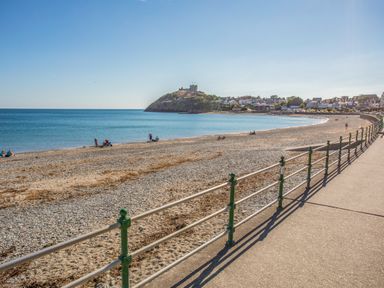 A beach with a stone promenade and railings near a coastal town with a hill and castle in the background at The Hideaway Pod in Rhoslan near Criccieth
