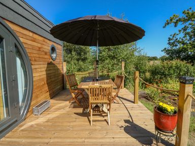 A wooden deck with a table, chairs, and a large umbrella outside a cabin at The Hideaway Pod in Rhoslan near Criccieth