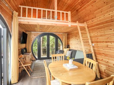 An interior of a wooden cabin with a dining table, chairs, a sofa, a ladder leading to a loft, and large circular doors at The Hideaway Pod in Rhoslan near Criccieth