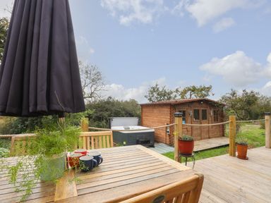 An outdoor wooden deck with a table, chairs, a potted plant, mugs, a closed umbrella, and a small wooden cabin near a hot tub at The Hideaway Pod in Rhoslan near Criccieth