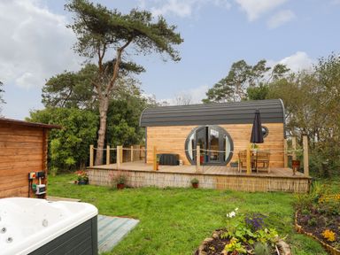 A wooden pod with circular windows on a raised deck with a table and chairs and a garden with a hot tub at The Hideaway Pod in Rhoslan near Criccieth