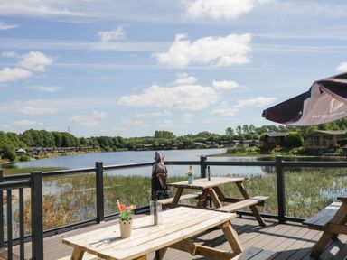 A wooden deck with picnic tables and flowers overlooking a lake with houses and trees at Parkers Place in Carnforth
