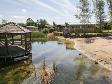 A lakeside with a wooden gazebo a sandy shore and several cabins in the background at Parkers Place Carnforth