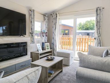 A living room with a television above a fireplace a wooden coffee table a small cabinet with vases and framed photos and sliding glass doors at Parkers Place in Carnforth