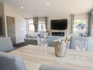 A dining table with grey chairs and a champagne bucket with glasses in a living room with sofas a television and windows at Parkers Place in Carnforth