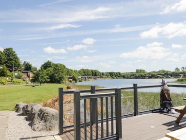 View of a lake with grass and trees alongside and wooden deck with railing in foreground at Parkers Place in Carnforth