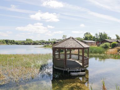 A wooden gazebo on water near reeds with houses and trees in the background at Parkers Place in Carnforth