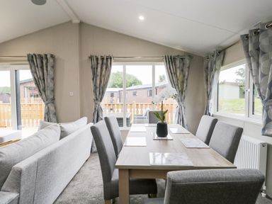 A dining area with a wooden table and six gray chairs next to windows with patterned curtains at Parkers Place in Carnforth