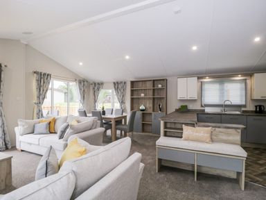 An open living room with two sofas and cushions a dining table with six chairs a kitchen with cabinets a sink and a bench with a cushion and a pillow at Parkers Place in Carnforth