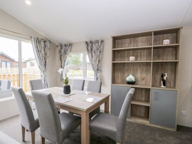 A dining area with a wooden table and six gray upholstered chairs near windows with patterned curtains and a wooden shelving unit with decorative items at Parkers Place in Carnforth